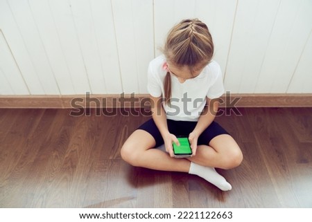 A girl who sits on the floor and uses a phone with a green screen. The child is sitting and looking into a smartphone with a colored key. High quality photo
