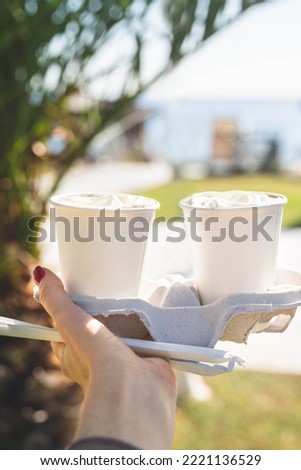 two paper cups in a glass holder held by a woman's hand against the backdrop of a palm tree and the sea. mockup