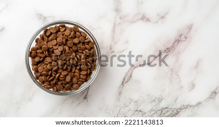 Dry pet food in a glass bowl over marble background. Glass feeding bowl full of gluten free dry protein kibbles for cats closeup. Complete food for domestic animals concept. Copy space. Top view. Royalty-Free Stock Photo #2221143813