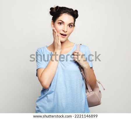 Charming young woman with dark hair in a blue t-shirt with a backpack looks surprised and joyful on a gray background. Good news.
