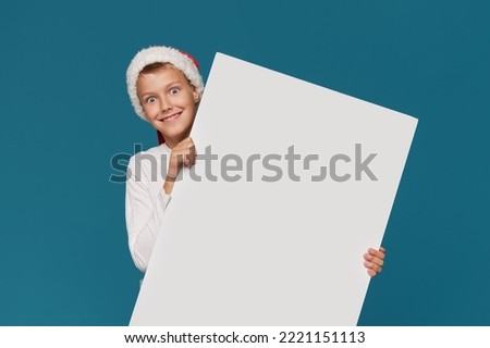 Surprised teen boy  in a Santa Claus hat and party glasses on a turquoise background. holds a white board sign for advertising, Empty space for text. wow funny face