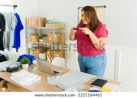 Young woman seller taking photos of t-shirts with her smartphone for her online shop and social media 