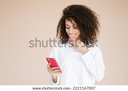 Adorable african american young woman holds mobile phone, browses social networks, has broad smile, enjoys online chatting, isolated on beige background, being always in touch                         