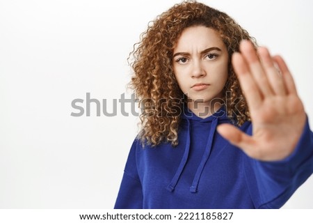 Portrait of young teen girl extending hand, showing stop, taboo prohibit gesture, disapprove smth, forbid, standing in blue hoodie over white background