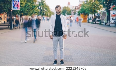 Portrait of young businessman in trendy garments standing in busy pedestrian street and looking at camera while crowds of people are whizzing around. Royalty-Free Stock Photo #2221248705