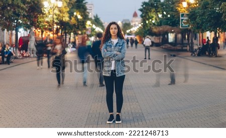 Portrait of stylish young lady tired of usual haste standing in the street among whizzing people and looking at camera. Time, youth and modern society concept. Royalty-Free Stock Photo #2221248713
