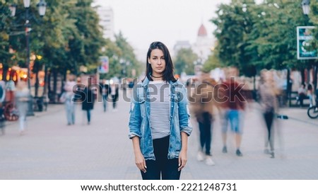 portrait of tired young woman student standing alone in city center and looking at camera with straight face while crowds of men and women are whizzing around. Royalty-Free Stock Photo #2221248731