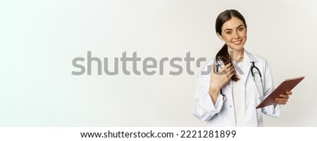 Smiling brunette female doctor, holding digital tablet, pointing at herself with friendly face, standing in white coat over white background.