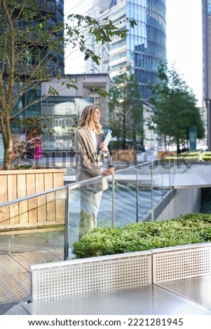 Vertical shot of smiling office girl in suit, drinks her coffee outdoors in city center, standing near office building, grabs takeaway.