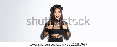 Image of happy satisfied asian woman dressed-up for halloween party, showing thumbs-up in approval, smiling pleased over white background. Girl in witch costume looking delighted.