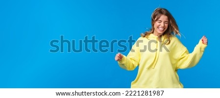 Girls just want ahve fun. Carefree and happy laughing blond girl enjoy spring, jumping and dancing as listening music on concert, shaking head as hair floating in air, blue background.