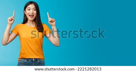 Amused asian girl introduce new product sharing promo with you, raise hands pointing fingers up smiling broadly, look excited and upbeat, enthusiastic advertising, stand blue background.