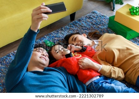 Happy Asian family dad using smartphone camera taking selfie photo with mom and daughter wear sweater lay down on carpet floor in living room celebrating Christmas eve and boxing day event at home.