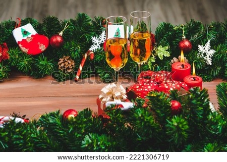 Lighten festive red candles, gift box and champagne glasses on wood bord, framed with green pine branches. Christmas and new year celebration concept.