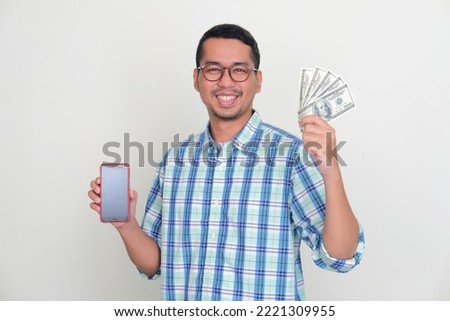 Adult Asian man smiling happy while holding US dollar currency and showing empty handphone screen