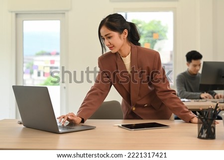 Attractive young female manager standing at her office desk and using laptop computer