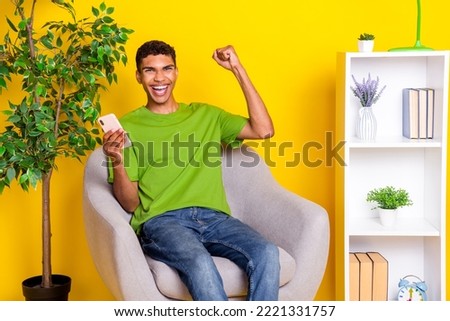 Photo of funny lucky guy wear green t-shirt sitting chair rising fist writing modern gadget isolated yellow color background
