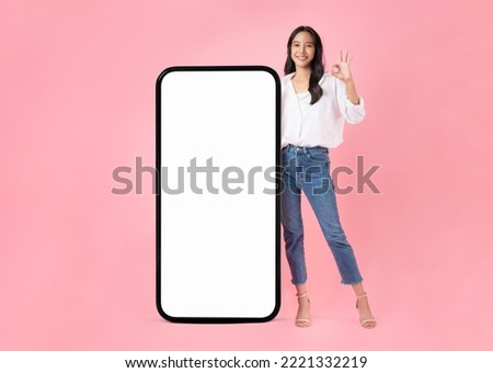 Beautiful Asian woman shows ok sign and stand with big smartphone mockup of blank screen and smiling on pink background.