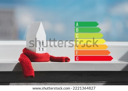 Miniature house in a red scarf and energy efficiency chart on the windowsill. The concept of passive house heating. Thermal insulation of a building or dwelling. Energy crisis. Royalty-Free Stock Photo #2221364827