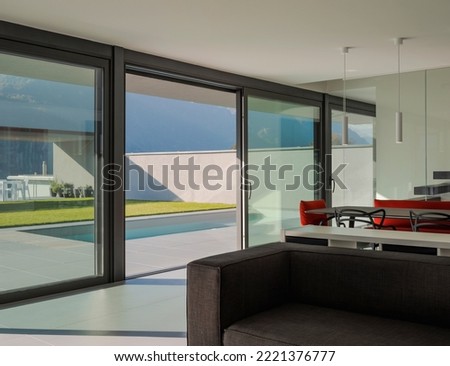 Interior modern villa living room with large finistre. Outside there is a swimming pool. Sunny day in the Alps of Switzerland. Nobody inside