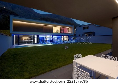 Modern house with swimming pool and garden in night scene illuminated by colored LED lights. Behind the house is the hill with the forest. Nobody inside