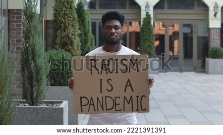 Young African American man stands with a cardboard poster RACISM IS A PANDEMIC in public outdoor place. An anti racist movement to protest against injustice and police brutality. Street demonstration.