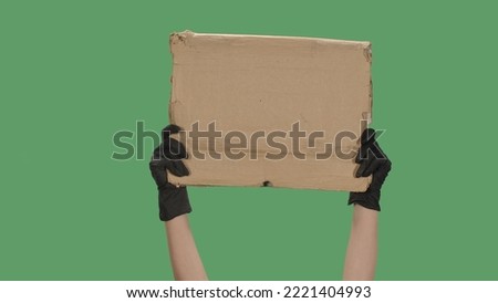 Hands in black gloves raise a blank poster made of a cardboard box. Empty space for your text, logo or advertisement. Banner design concept. Isolated a green screen, chroma key.