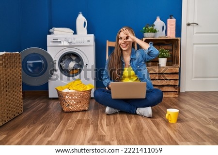 Young blonde woman doing laundry using laptop smiling happy doing ok sign with hand on eye looking through fingers 