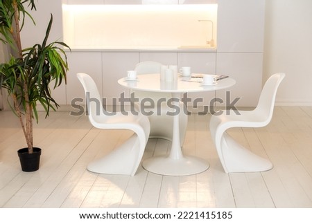 Modern Kitchen interior design with furniture. Stylish white dining room with large round table and designer chairs, cups coffee, large houseplant in pot. White plastic chairs pantone. 