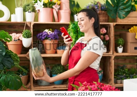 Young beautiful hispanic woman florist make photo by smartphone to plant jar at flower shop