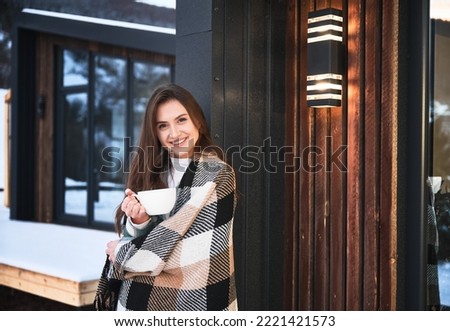 Young woman resting on terrace of modern barn house in the mountains. Happy female tourist holding cup of tea, enjoying winter vacation in new cottage.