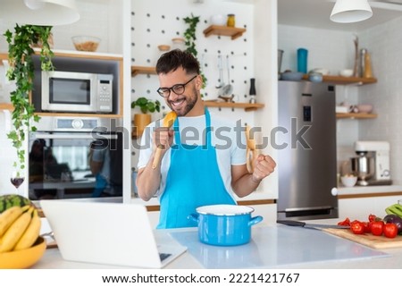 Funny young man singing song into whisk, cooking in modern kitchen, happy male holding kitchenware as microphone, listening to music, dancing, doing housework at home, preparing breakfast