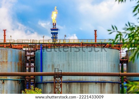 A burning gas torch against a blue sky against the background of fuel tanks at a factory. Gas processing and oil refining. Exhaust gases, gas crisis, greenhouse effect.