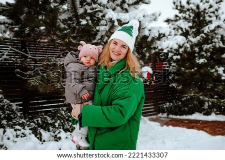 Pretty young woman in green winter coat is playing with her baby girl outside while snowing time. Winter concept.  Royalty-Free Stock Photo #2221433307