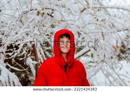 Portrait of cute teen boy in red winter jacket against snowed winter garden background.