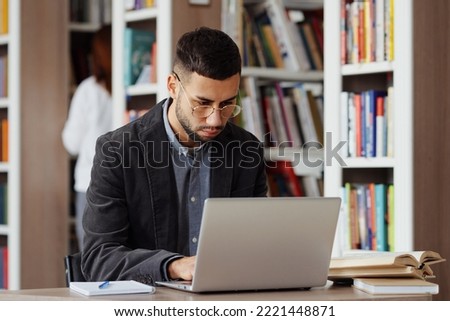 Intelligent man in eyeglasses wearing jacket working on laptop in library, books lying beside, researcher browsing internet, student doing assignment, person on blurred background choosing book