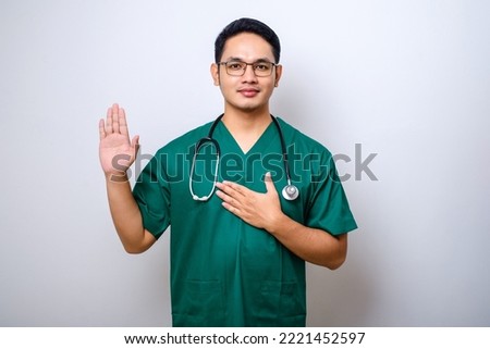 Smiling honest asian male nurse or doctor, intern in scrubs giving pledge, raising one arm and hold hand on heart while promise, oath to patient over white background Royalty-Free Stock Photo #2221452597