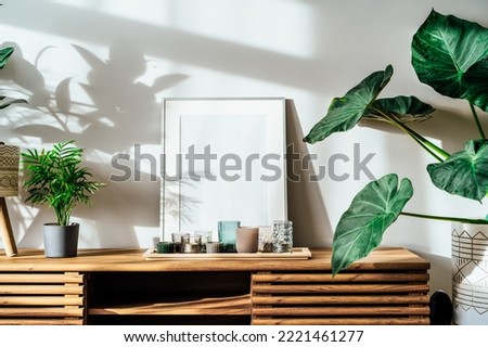 Modern minimalist Scandinavian style interior with white poster mockup, candles and tropical green home plants on a wooden console under sunlight and shadows on a gray wall. Selective focus.