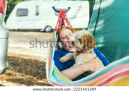 Relaxed woman listening to music with headphones lying in hammock with cockapoo puppy near motorhome on camping trip. Female living on camper car with animal, travel the world. Caravan car Vacation Royalty-Free Stock Photo #2221461289
