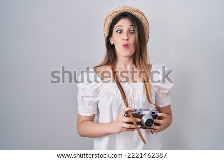 Young brunette woman holding vintage camera making fish face with mouth and squinting eyes, and comical. 