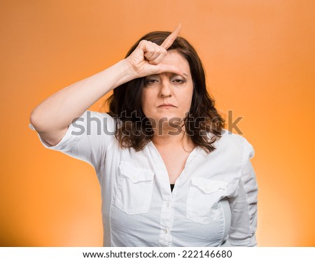 Loser. Closeup portrait funny mature middle aged woman giving loser sign on forehead, looking at you disgust on face isolated orange background. Negative human emotion facial expression body language