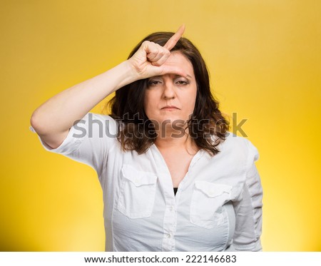 Loser. Closeup portrait funny mature middle aged woman giving loser sign on forehead, looking at you disgust on face isolated yellow background. Negative human emotion facial expression body language