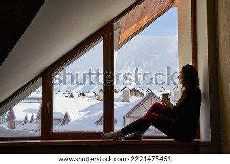 Woman being cold sits on windowsill, warming up with tea mug, looking at snow covered mountains. Female drinks coffee in cozy chalet wearing thermal underwear layer. Rising heating bills on winter. Royalty-Free Stock Photo #2221475451