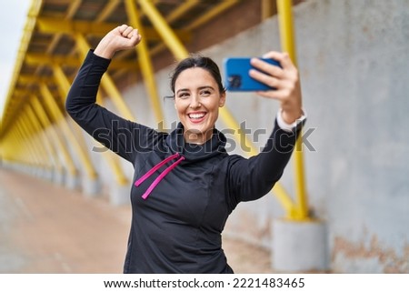 Young hispanic woman doing strong gesture making selfie by the smartphone at street