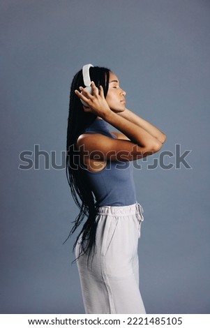 Sideview of a girl with braids listening to music on wireless headphones in a studio. Young woman enjoying her favourite audio playlist while standing against a grey background.
