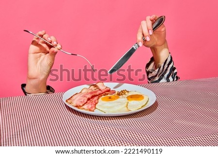 Female hands with fork and knife eating English breakfast with fried eggs and bacon . Vintage, retro style interior. Food pop art photography. Complementary colors. Copy space for ad, text Royalty-Free Stock Photo #2221490119