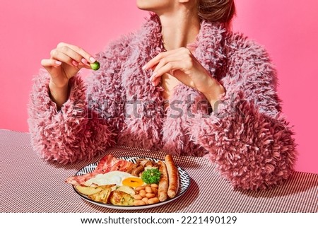 Woman in furry pink coat eating English breakfast with eggs, bacon, sausage and vegetables. Vintage, retro style interior. Food pop art photography. Complementary colors. Copy space for ad, text Royalty-Free Stock Photo #2221490129