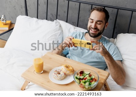 Young man wearing blue t-shirt lying in bed have breakfast take picture shot on mobile cell phone of food rest relax spend time in bedroom lounge home in own room house. Good mood bedtime concept.