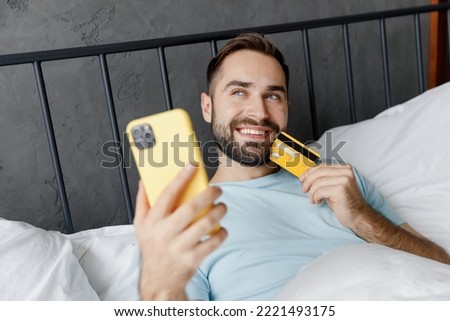 Young minded man in blue t-shirt lying in bed using mobile cell phone hold credit bank card shopping online book tour rest relax spend time in bedroom home in own room. Good mood night bedtime concept