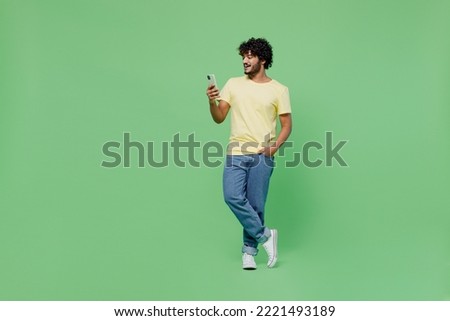 Full body cool young smiling happy Indian man 20s in basic yellow t-shirt hold in hand use mobile cell phone isolated on plain pastel light green background studio portrait. People lifestyle concept